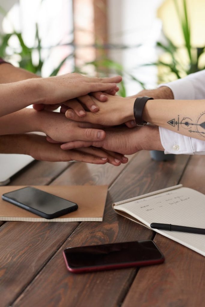 photo-of-people-near-wooden-table-3184431 Hands from a diverse team stack on a table symbolizing unity and teamwork in a modern office setting.